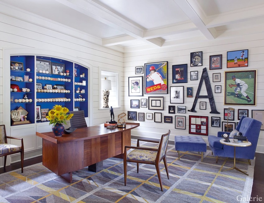 Home office with wooden desk, shelves of baseball memorabilia, sports-themed wall art, blue chairs, and sunflowers in vase.