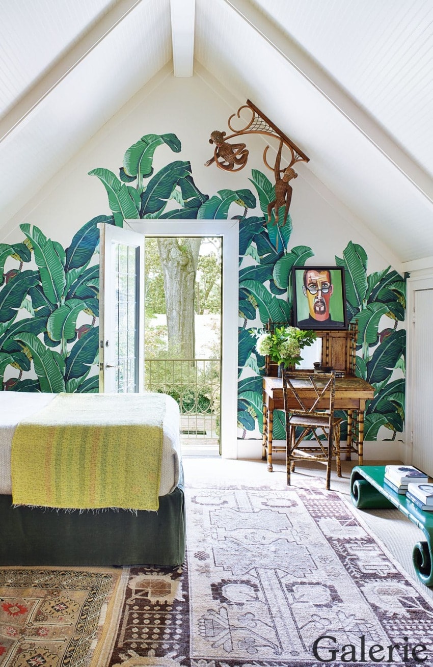 Attic bedroom with tropical wallpaper, natural light through open door, and eclectic furnishings including desk and artwork.