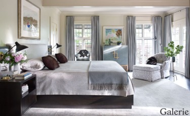 Elegant bedroom with king-size bed, decorative pillows, side tables, large windows, artwork, and an upholstered chair.