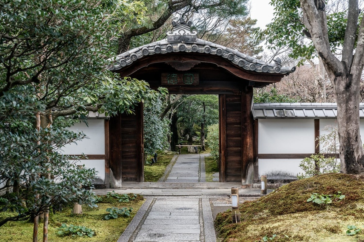 Traditional Japanese garden entrance with wooden gate, stone pathway, lush greenery, and trees in the background.
