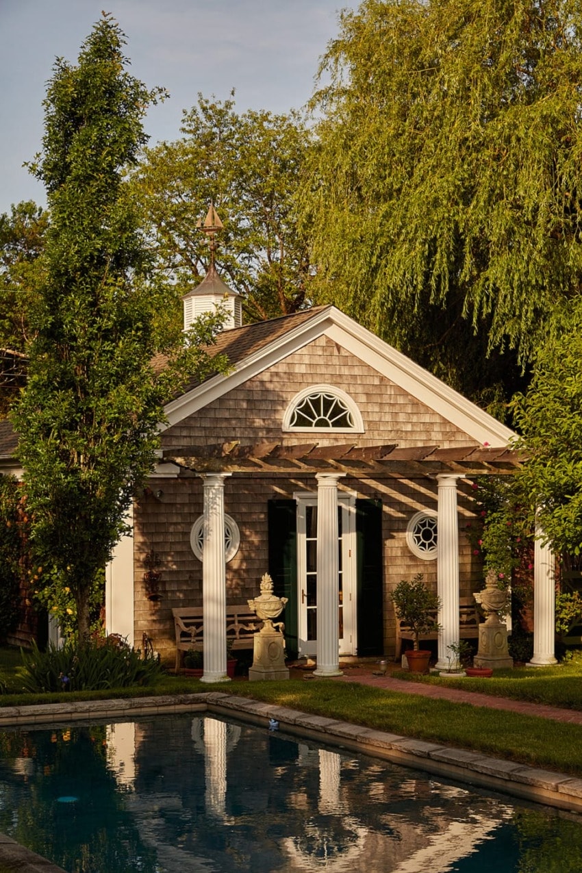 Charming cottage with pergola and pool surrounded by lush greenery and trees.