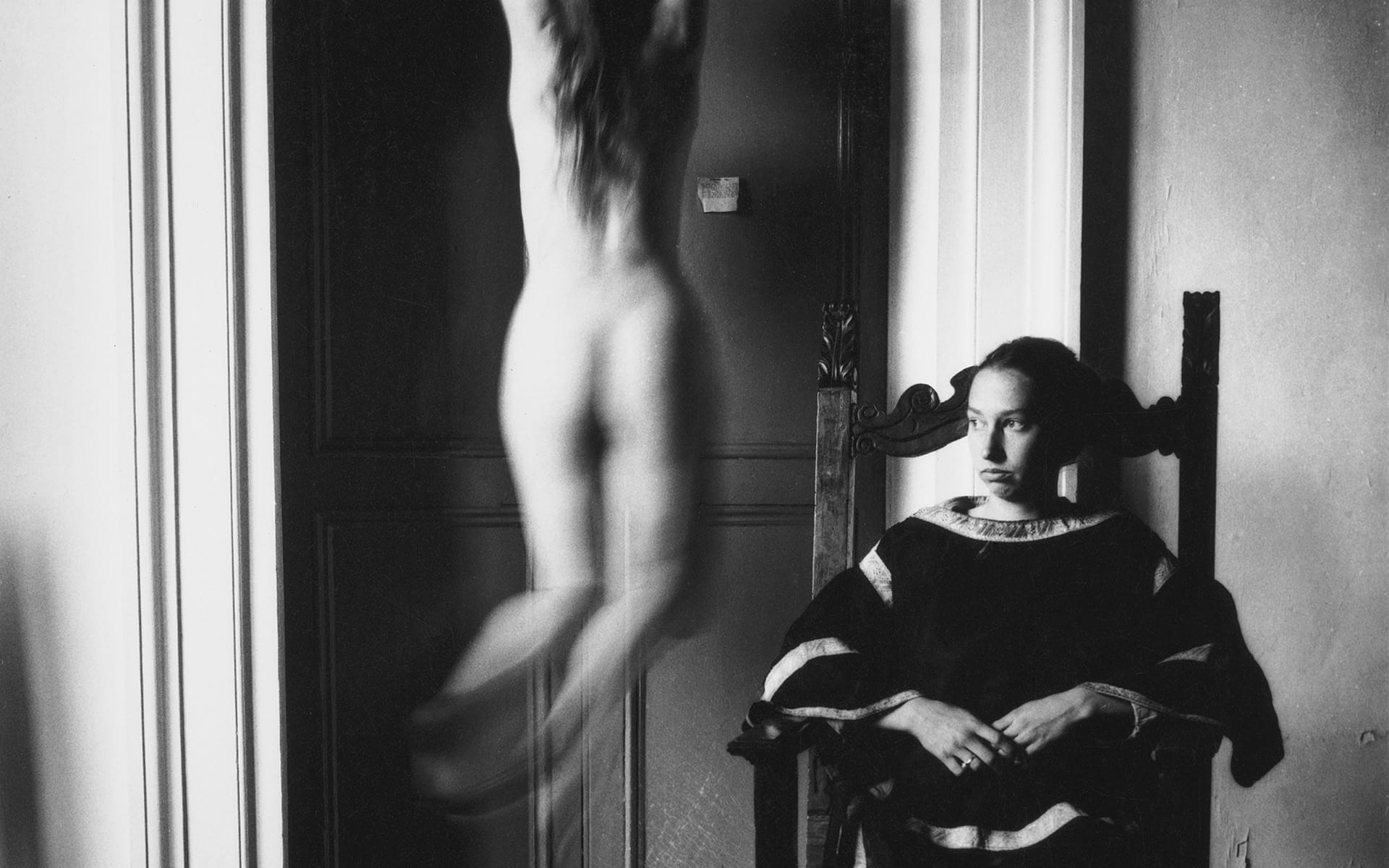 Person in dark clothing sits on ornate chair while blurred figure moves nearby in black and white photograph.