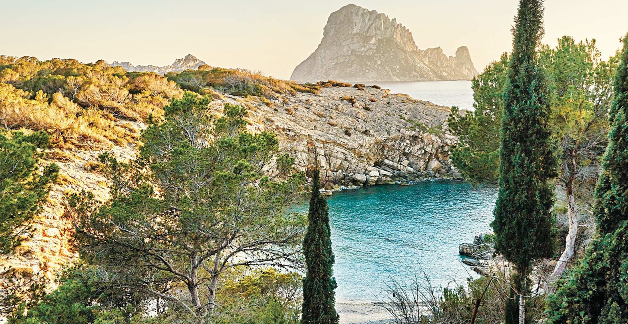 Scenic view of rocky coastline with turquoise water, lush greenery, and distant rock formation under a clear sky.