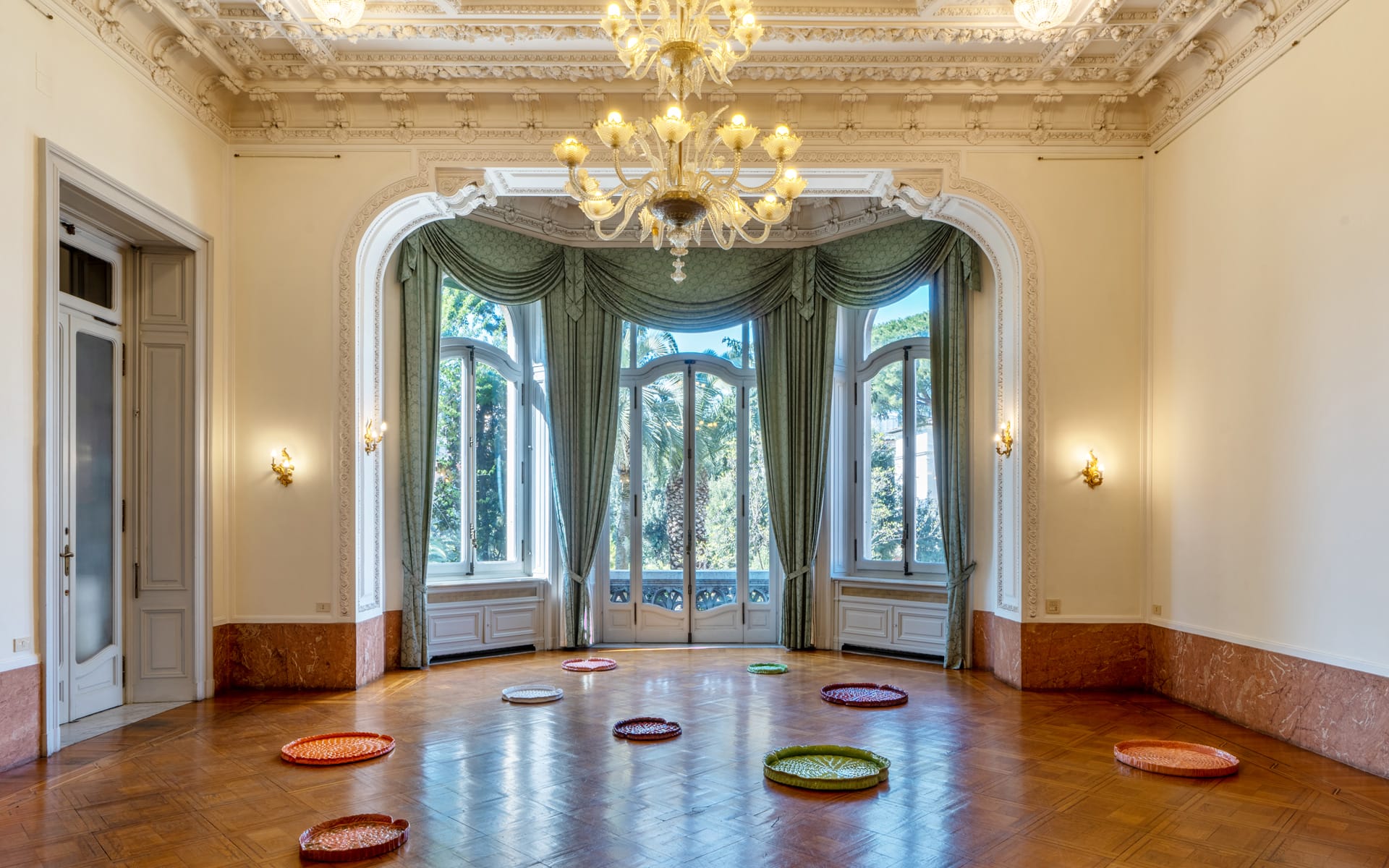 Elegant room with high ceilings, large arched windows, a grand chandelier, and colorful circular cushions on the wooden floor.