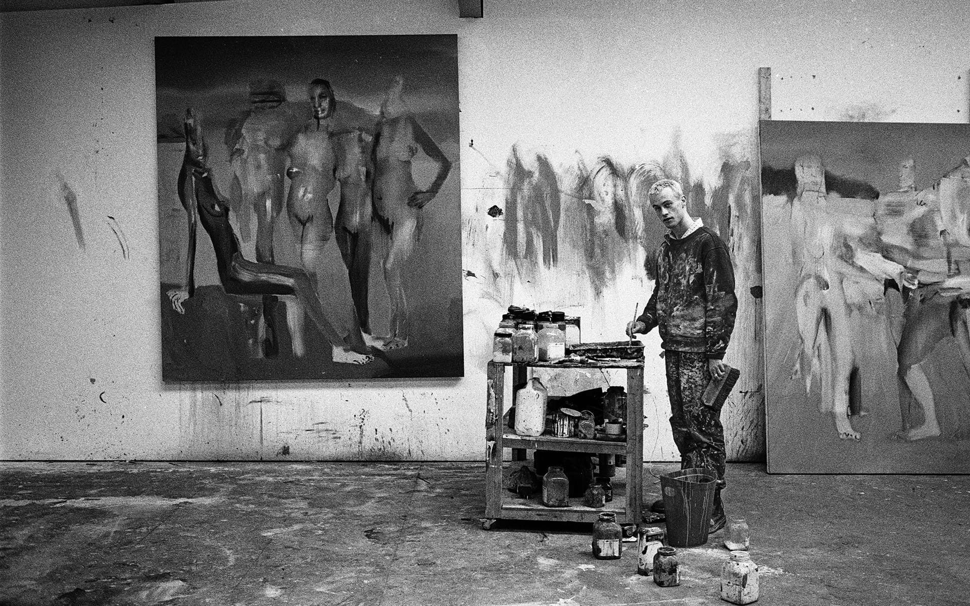 Artist in a studio with abstract paintings, standing next to a cart with art supplies, black and white photo.