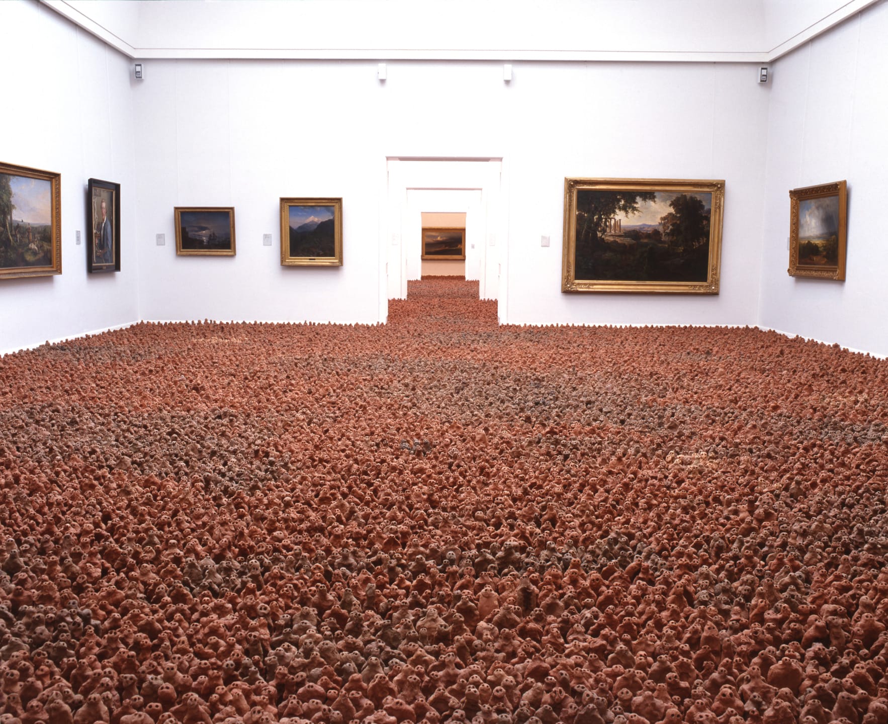 Art installation in a gallery with a floor covered in clay figures and landscape paintings on the walls.
