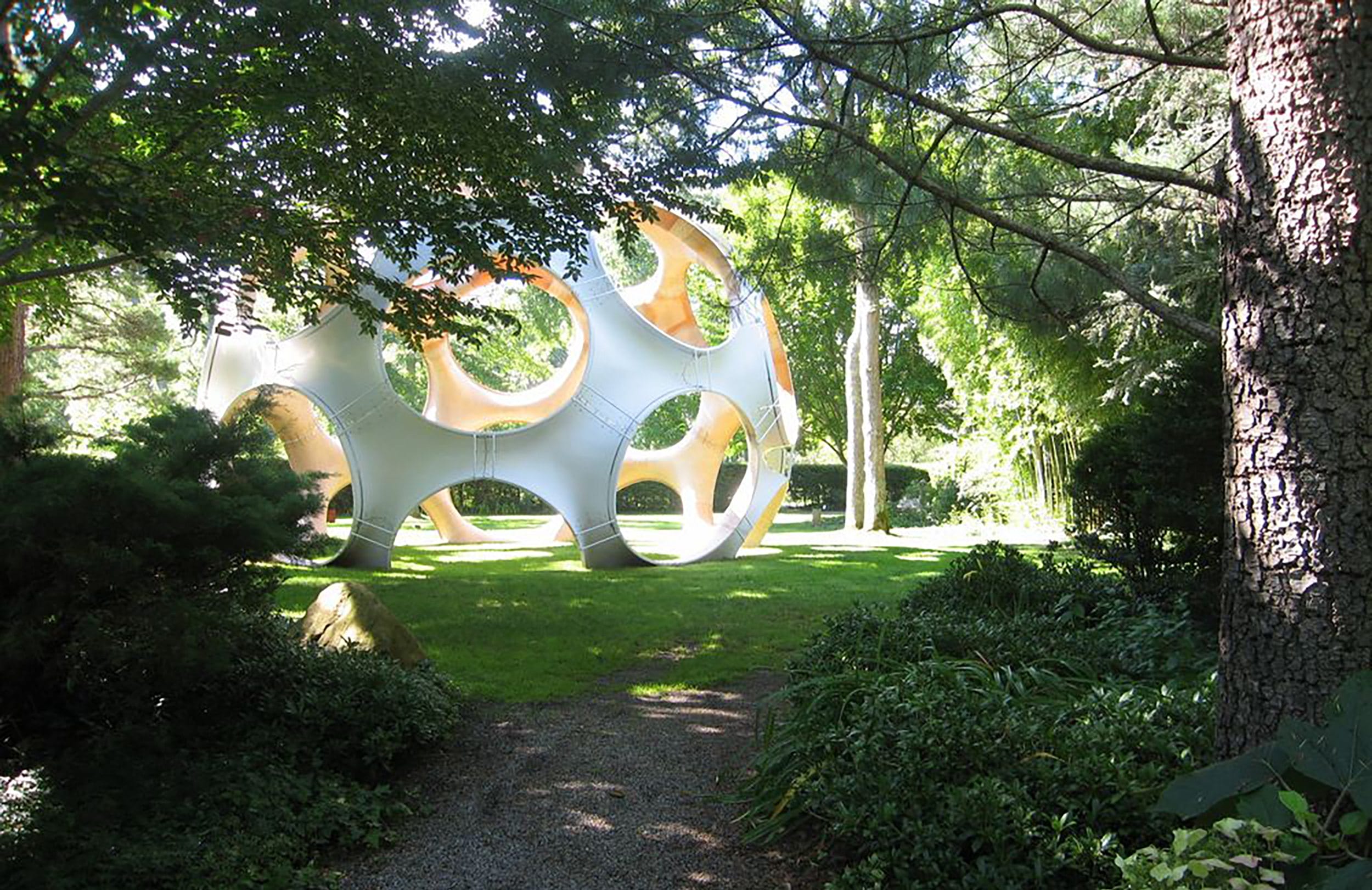 Sculptural structure in a lush green park, with sunlight filtering through the trees and casting shadows on the grass.