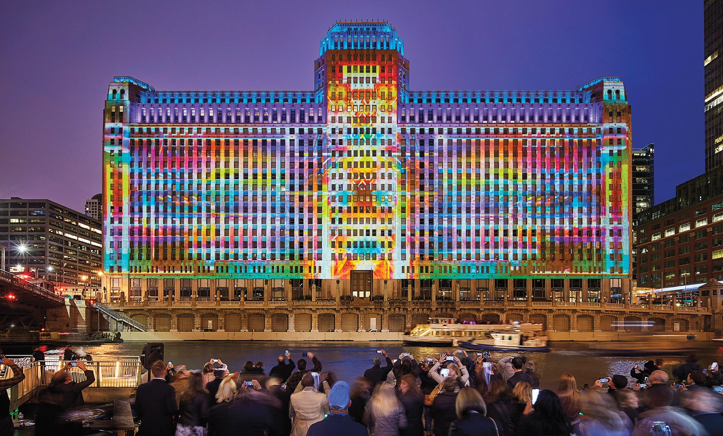 Colorful light projection on large building with people viewing from nearby riverbank at night.