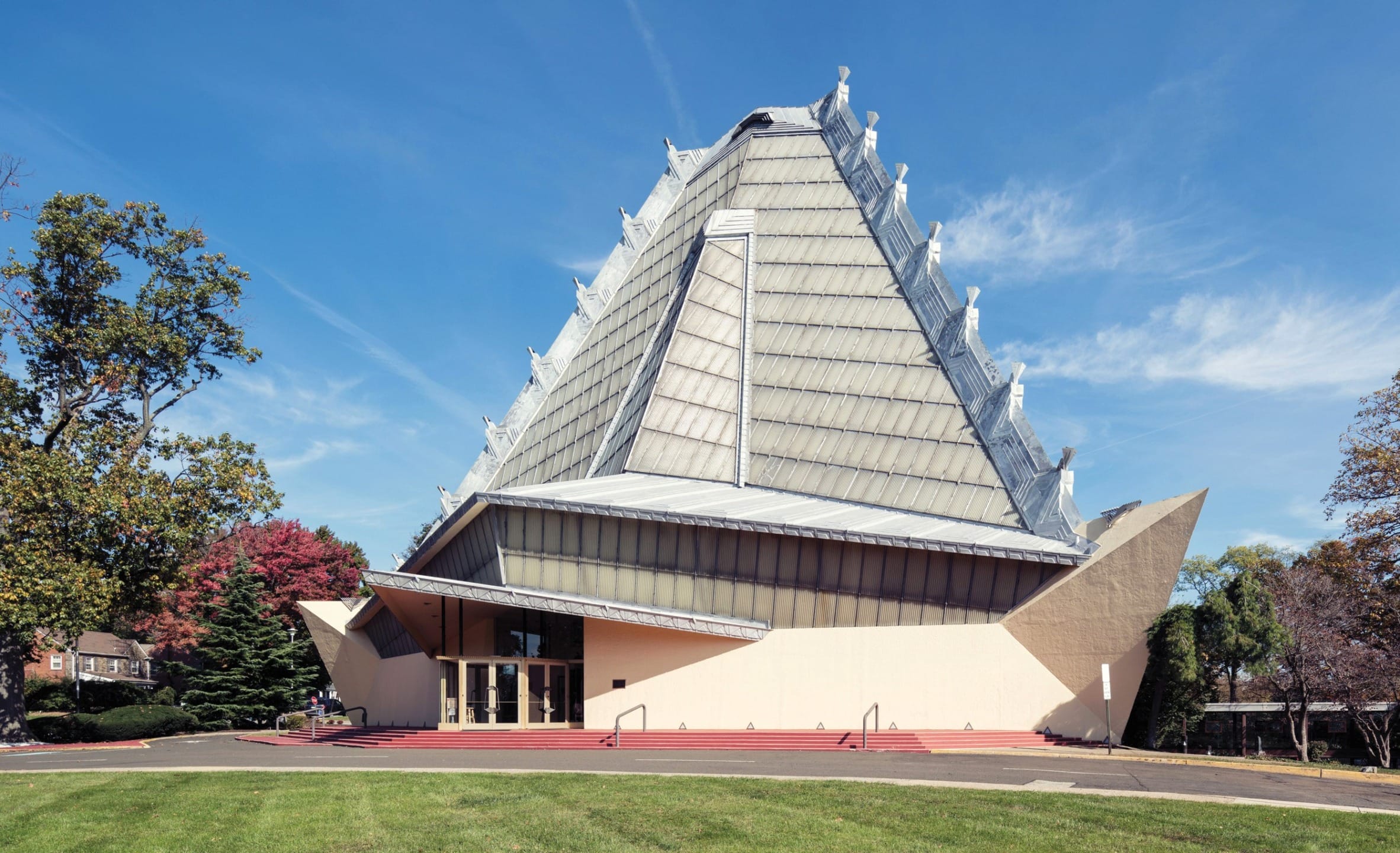 Modern triangular building with a steep, pointed roof, surrounded by trees on a sunny day.