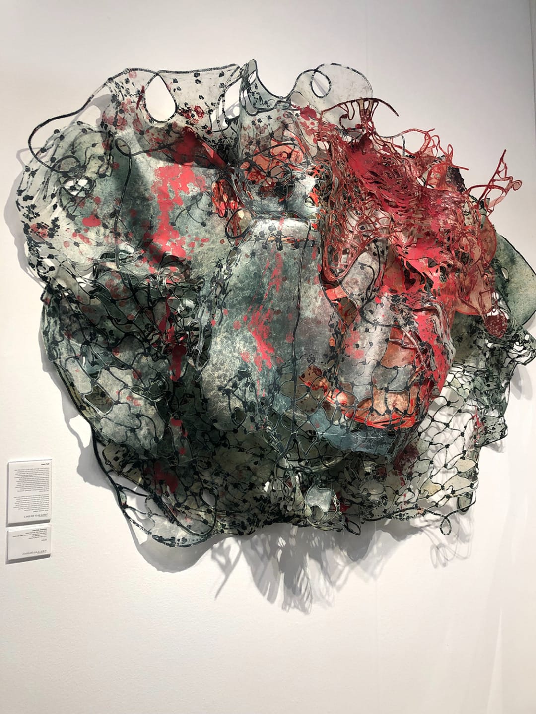 Abstract sculpture with intricate mesh design, featuring red and dark green colors, mounted on a white wall.