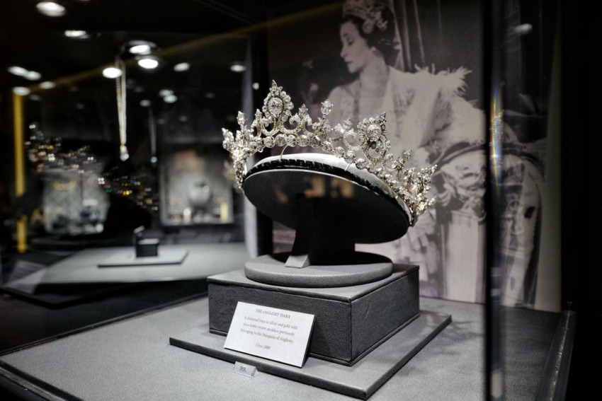 Ornate diamond tiara displayed in glass case, with a historical photograph in the background.
