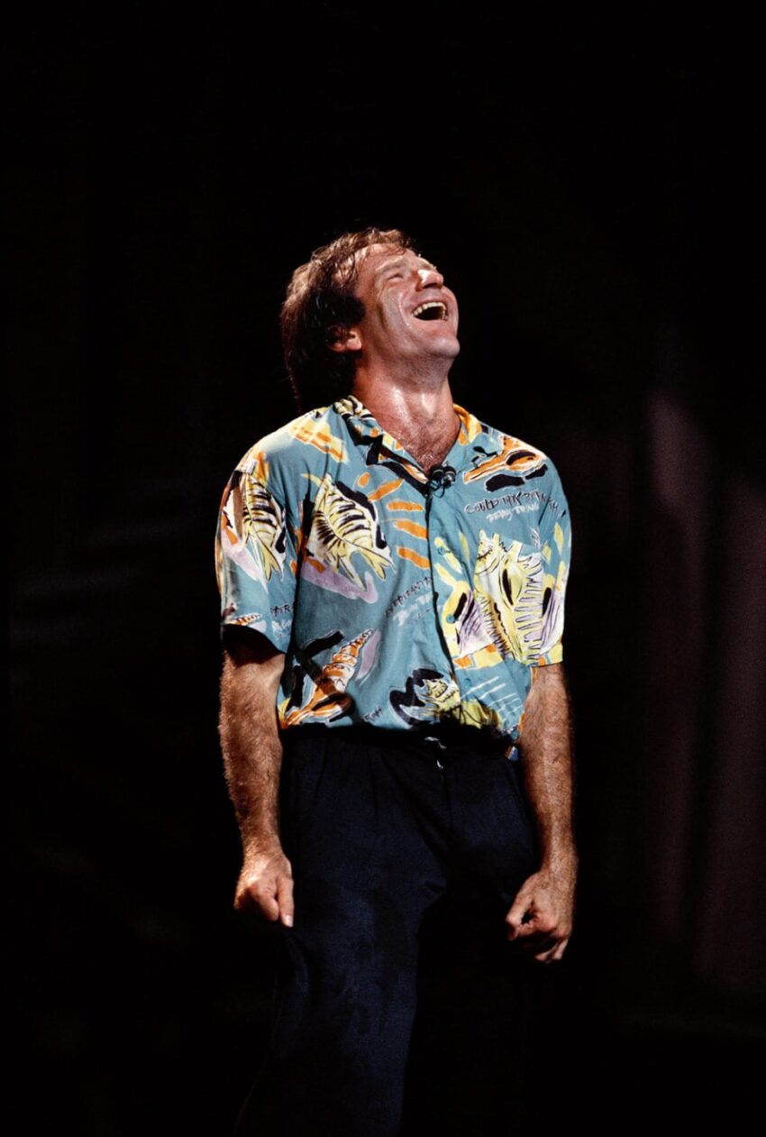 Man in a colorful shirt laughing heartily on stage against a dark background.