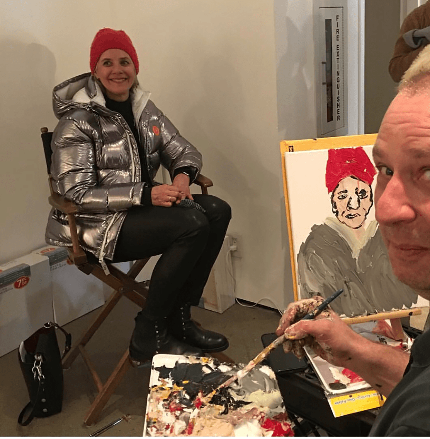 Woman in silver jacket and red hat poses for portrait while artist paints her image in a studio setting.