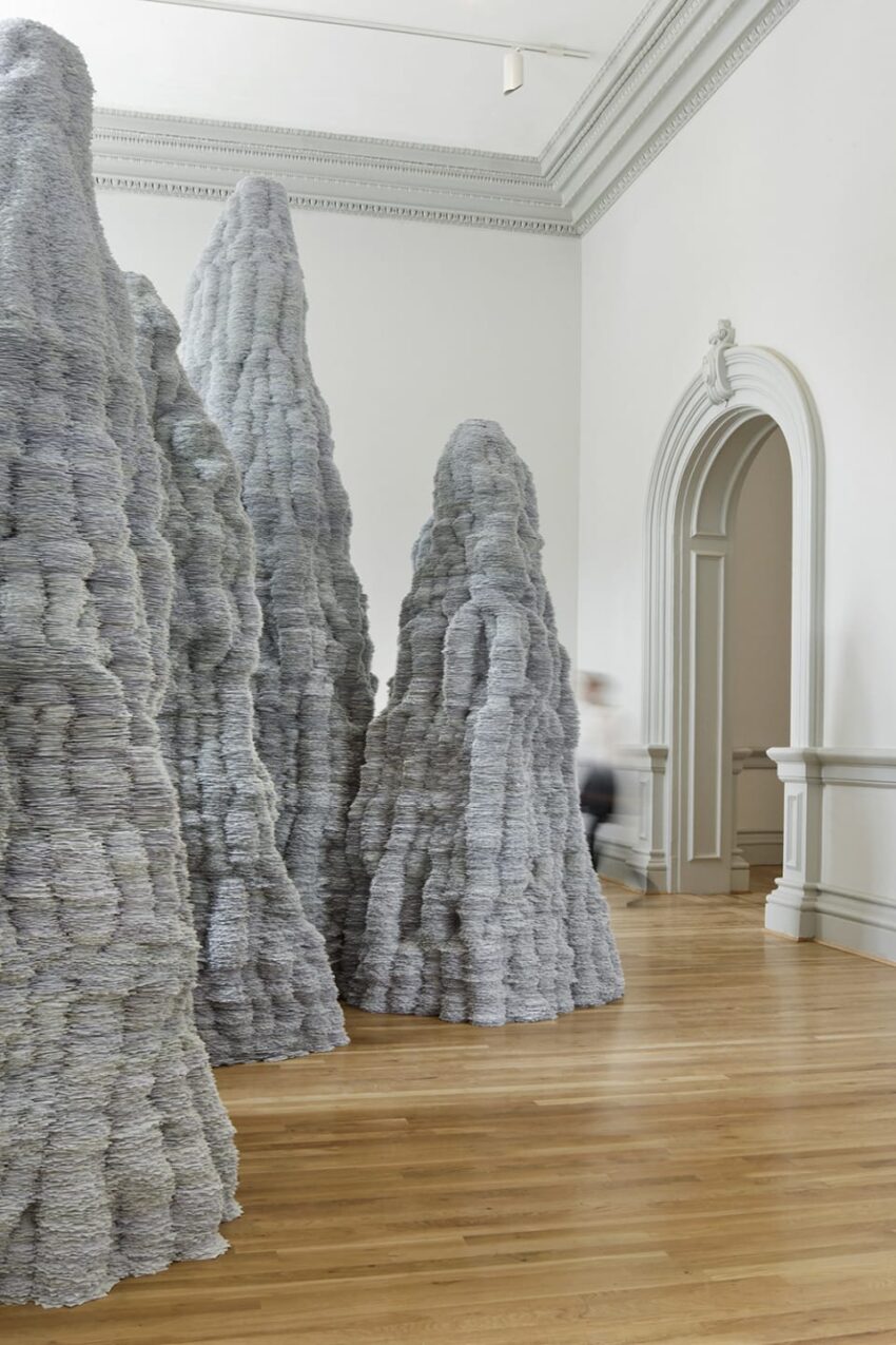 Tall, intricate sculptures resembling gray paper mountains displayed in a modern gallery with wooden floors and white walls.