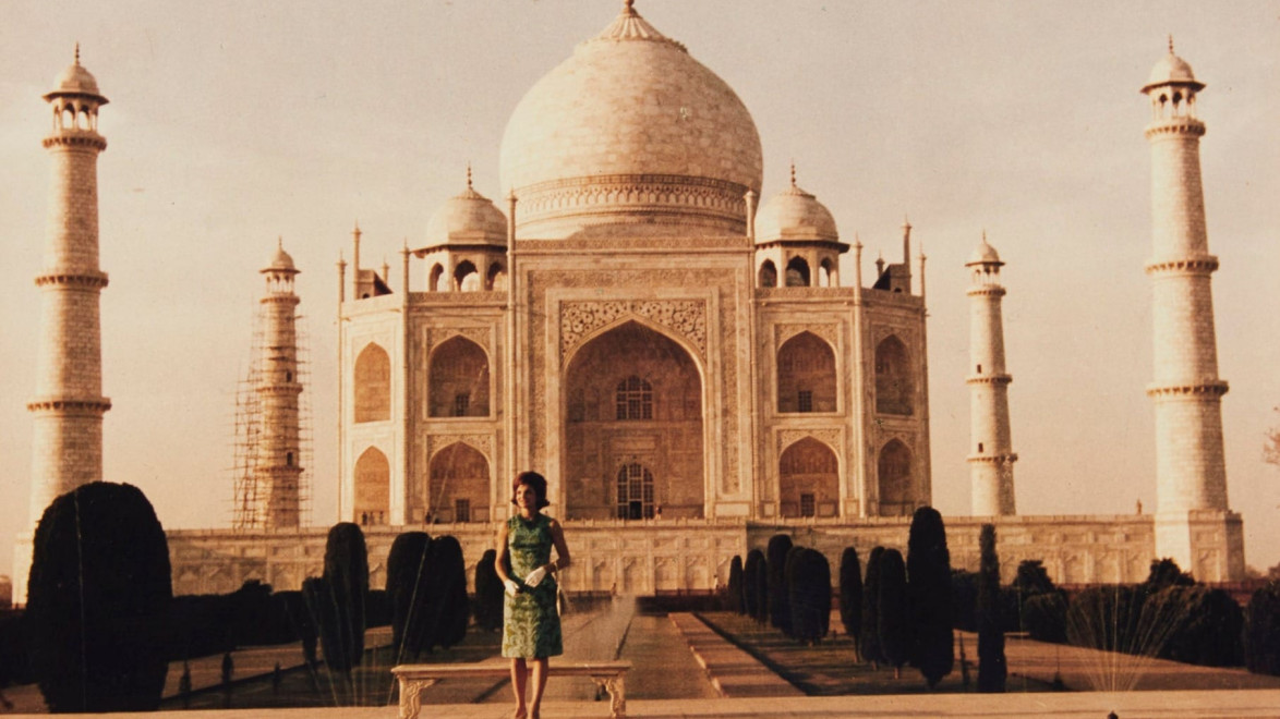 Woman in a green dress poses in front of the Taj Mahal with its reflection in the water on a clear day.