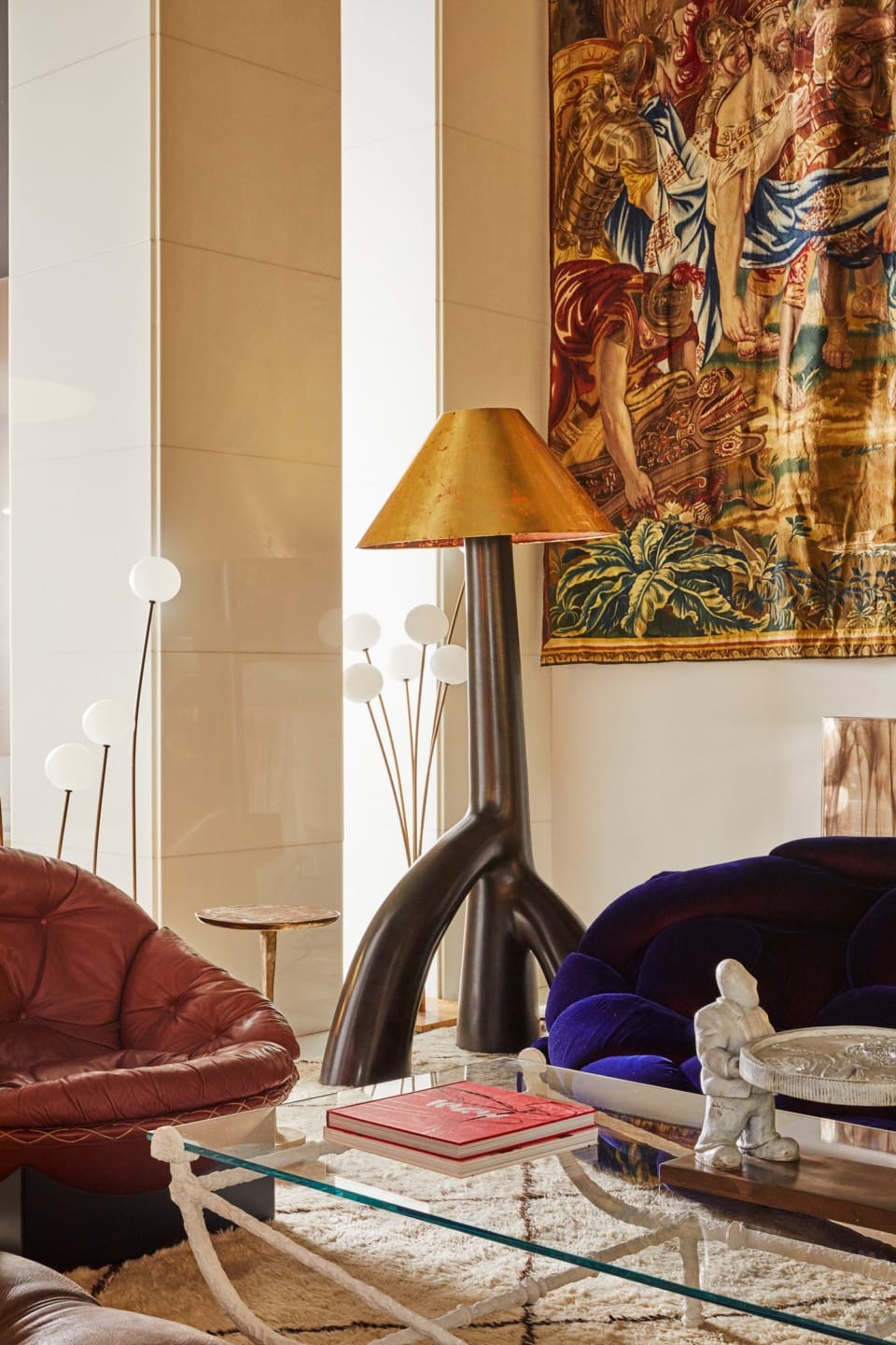 Modern living room with eclectic furniture, tall lamp, tapestry on the wall, and glass coffee table with books and sculpture.