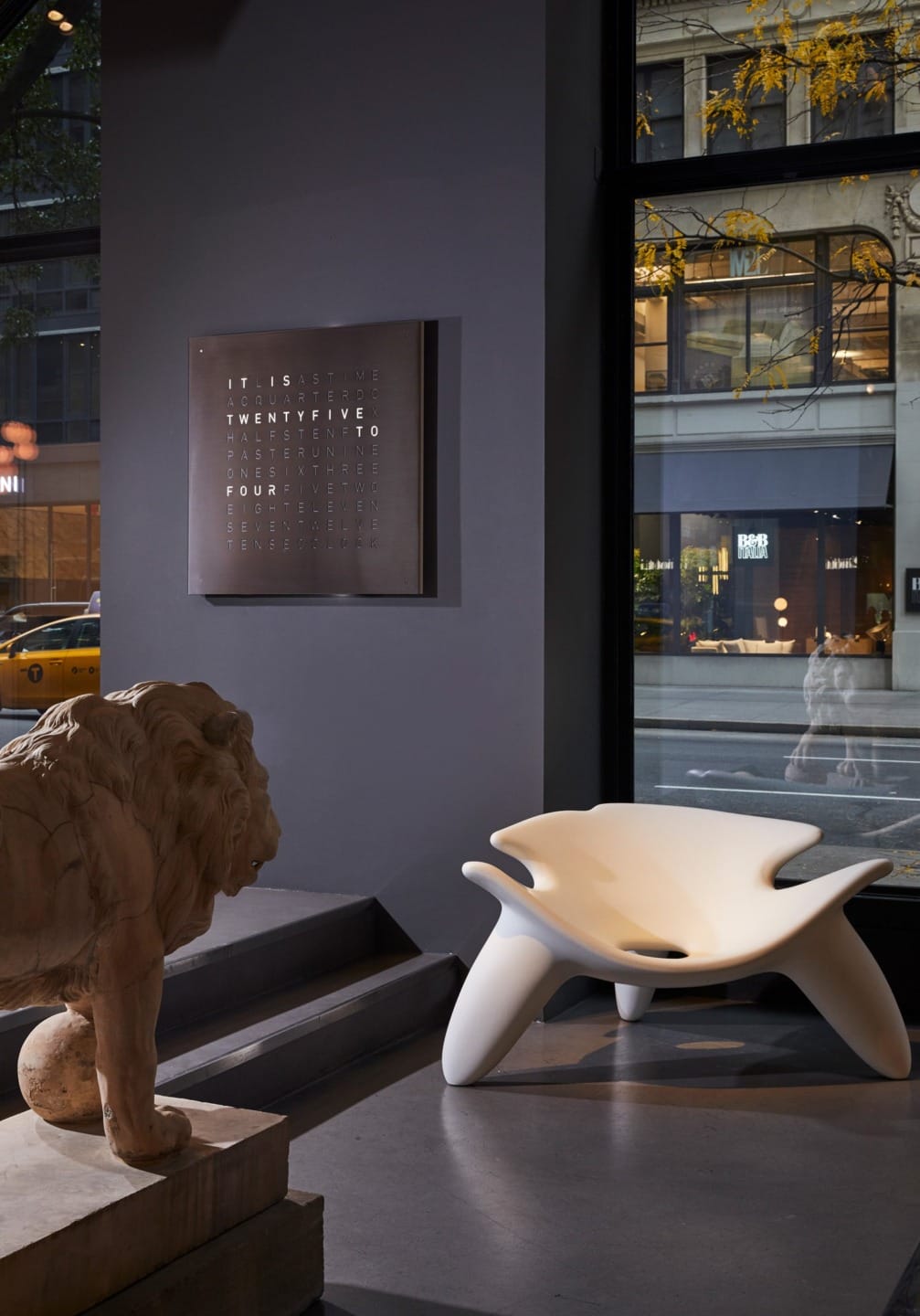 Modern indoor lounge area with a unique white chair, a lion sculpture, and a word clock on a dark wall beside a large window.