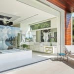 Luxurious modern bathroom with glass shower, bathtub, large windows, and a lounge chair on sunny balcony with greenery.