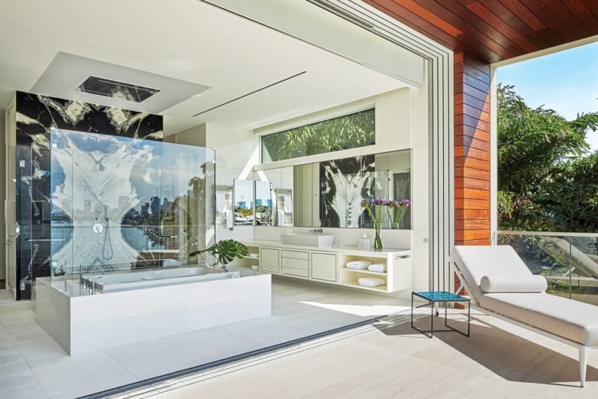 Luxurious modern bathroom with glass shower, bathtub, large windows, and a lounge chair on sunny balcony with greenery.