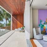 Modern balcony with large colorful abstract painting, wooden ceiling, cozy chairs, and tropical palm tree view.