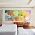 Modern bedroom with colorful abstract art, textured bedding, wooden dresser, and a view of a sunny balcony and greenery.