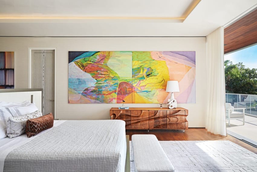 Modern bedroom with colorful abstract art, textured bedding, wooden dresser, and a view of a sunny balcony and greenery.
