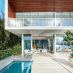 Modern two-story home with glass facade, pool, and outdoor seating area under a clear blue sky.