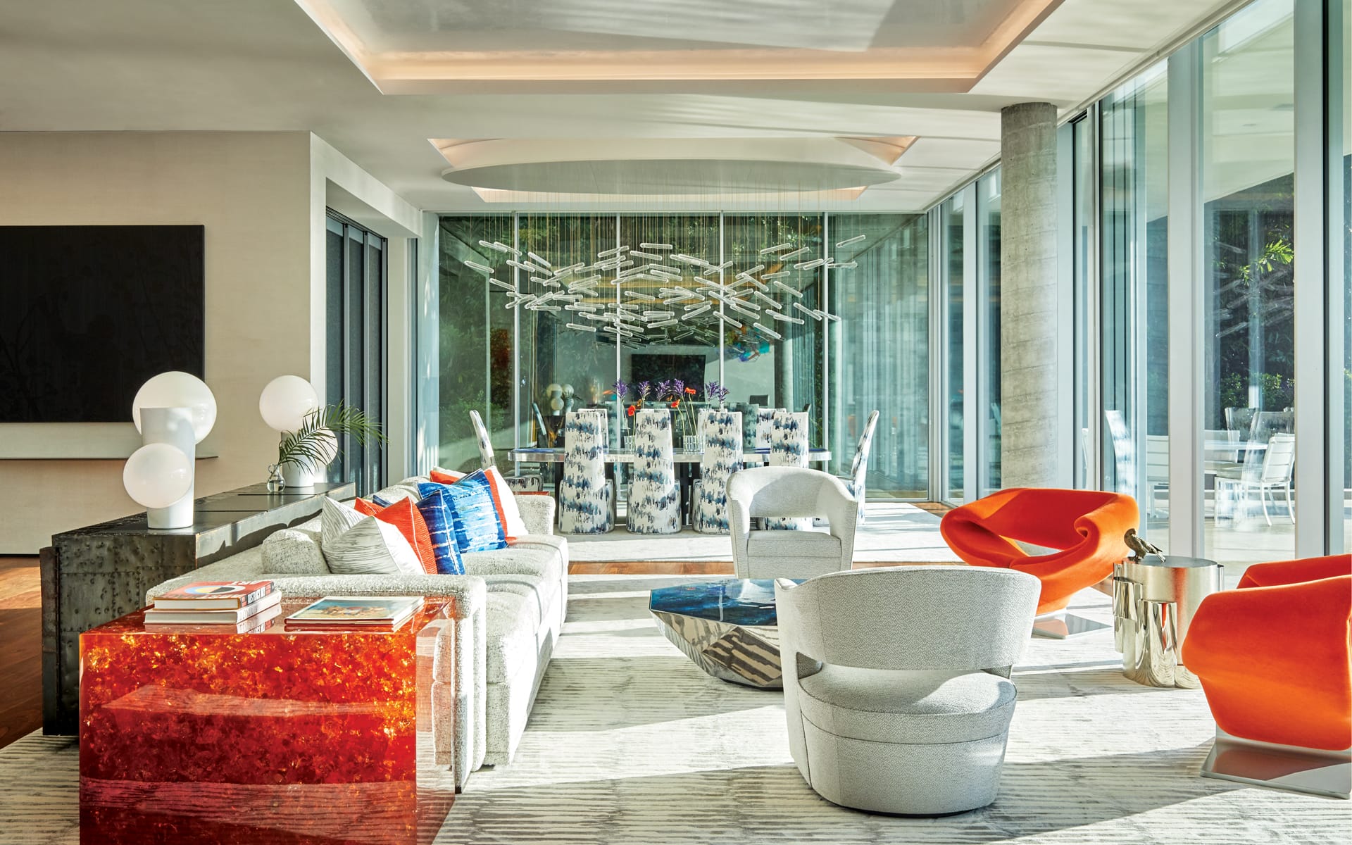 Modern living room with bright orange chairs, white sofas, glass walls, and abstract chandelier above a dining table.