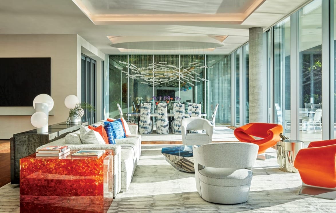 Modern living room with colorful furniture, large windows, abstract art, and a stylish dining area in the background.