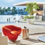 Modern living room with an orange chair overlooking a waterfront patio, large potted plant, and city skyline in background.