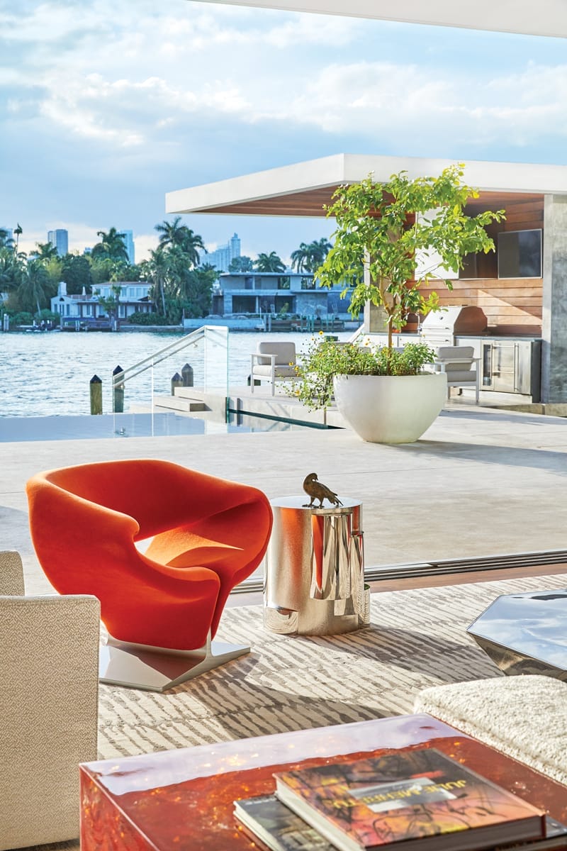 Modern living room with an orange chair overlooking a waterfront patio, large potted plant, and city skyline in background.