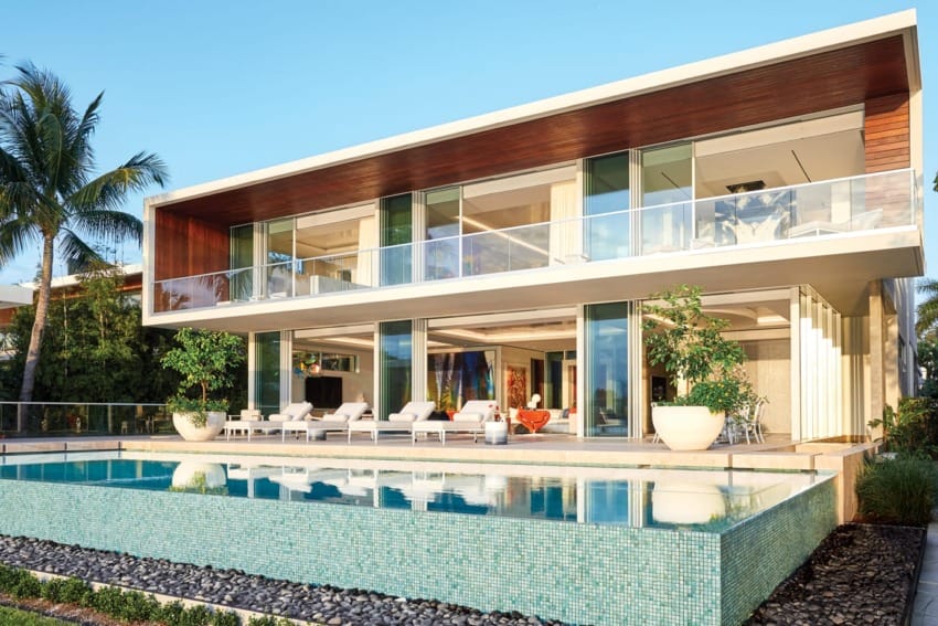 Modern luxury house with large glass windows, pool in foreground, surrounded by lush greenery and palm trees, under clear sky.