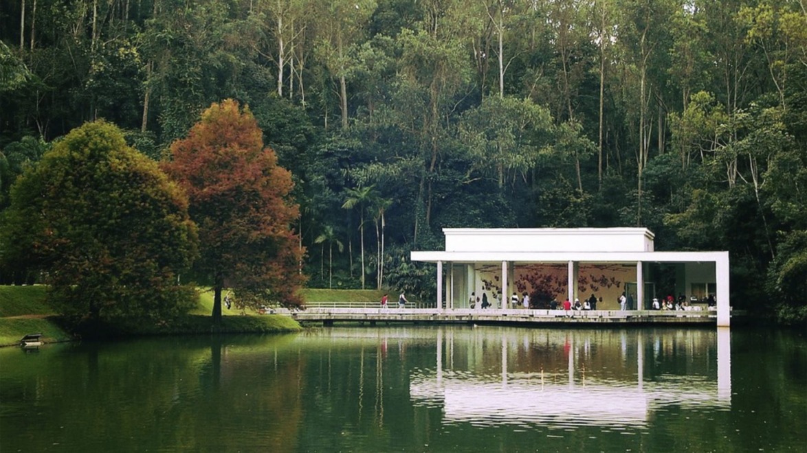 Modern white pavilion surrounded by lush trees reflected in a lake, with people inside, creating a serene natural setting.