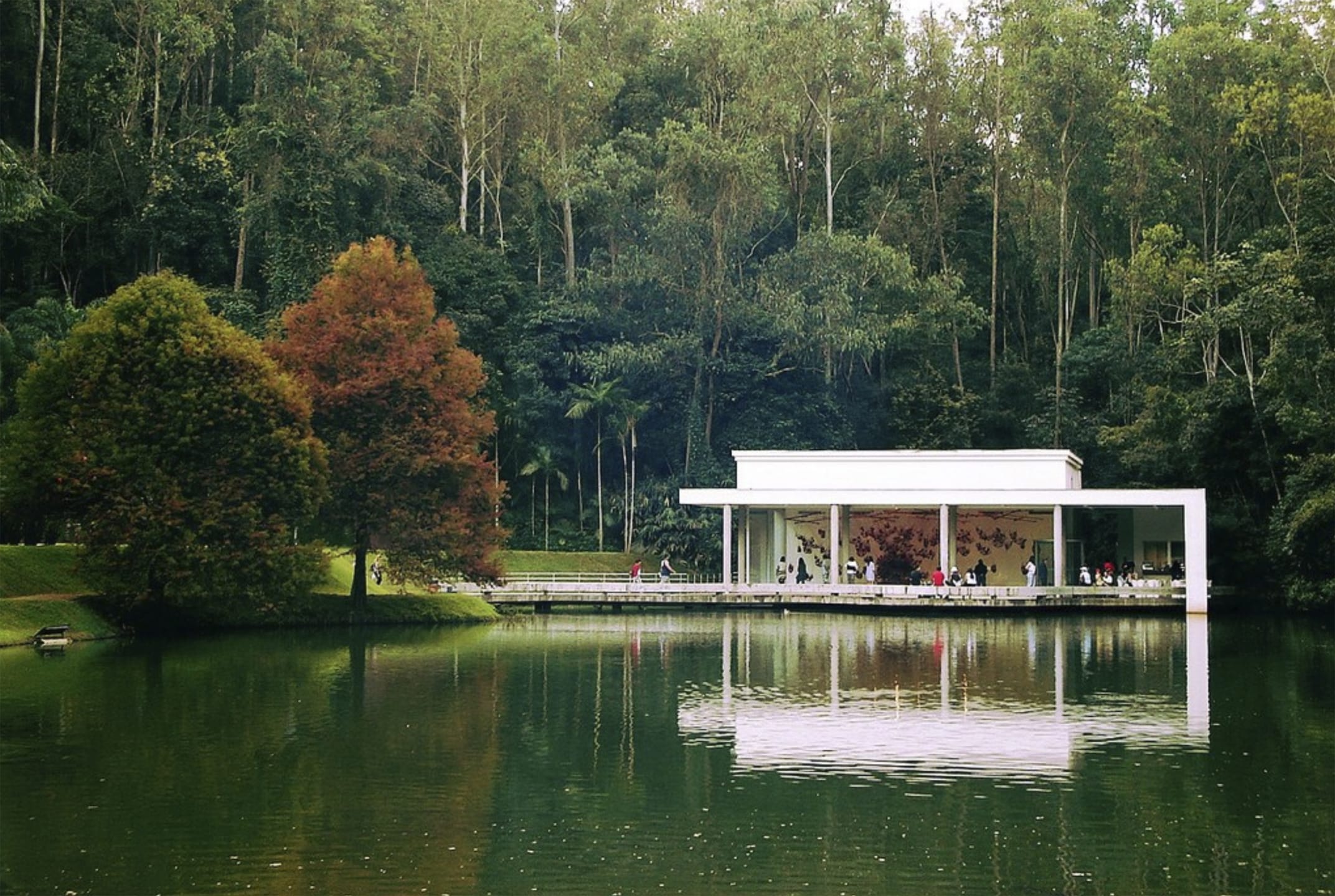 Modern white pavilion surrounded by lush trees reflected in a lake, with people inside, creating a serene natural setting.