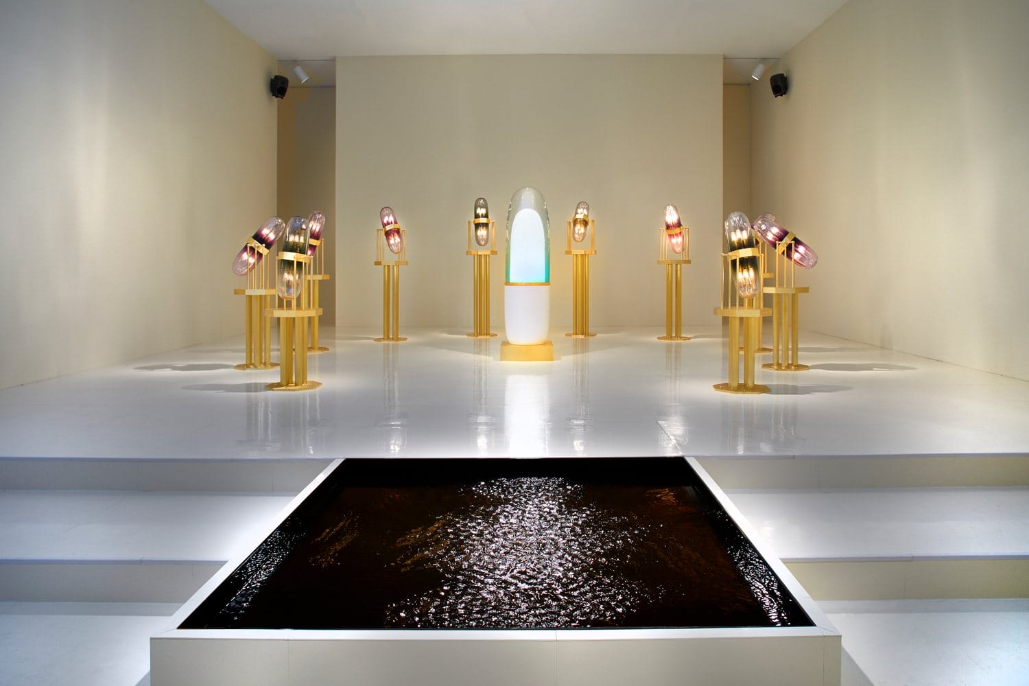 Modern art installation with illuminated sculptures on golden stands surrounding a central piece in a minimalist gallery space.