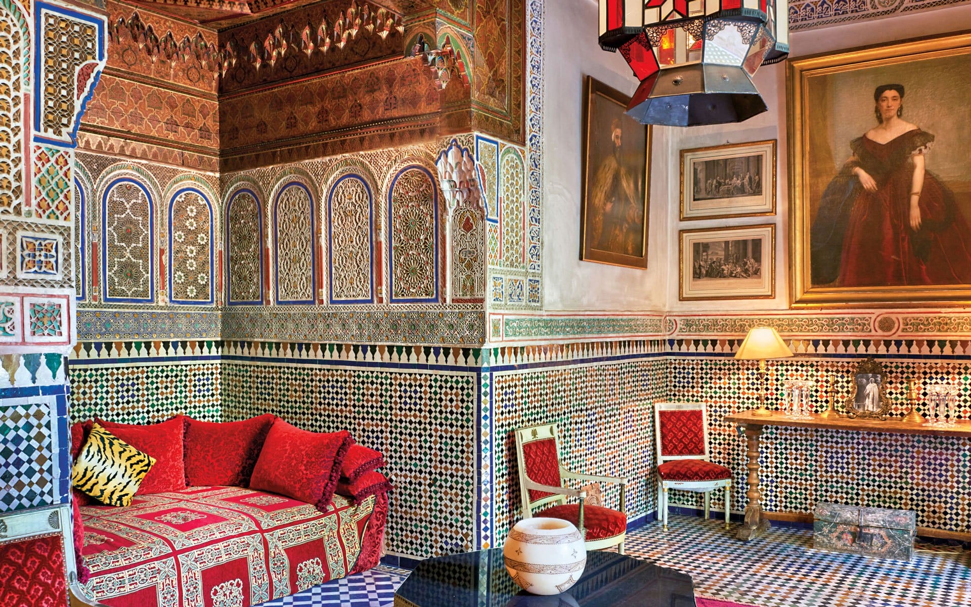 Ornate Moroccan room with patterned tiles, red sofa, artwork, and a hanging lamp.