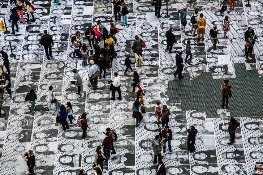 Crowd walking on pavement covered in black and white portraits, photographed from above.