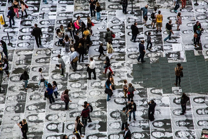 Crowd walking on pavement covered in black and white portraits, photographed from above.