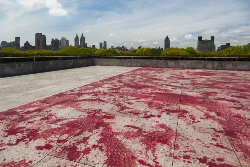 Rooftop art installation with red paint on concrete, overlooking city skyline and green trees under a partly cloudy sky.