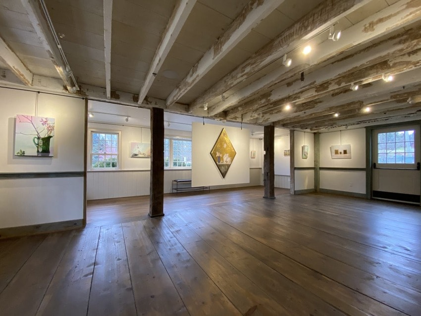Art gallery interior with wooden beams, showcasing various paintings on white walls, under soft lighting.
