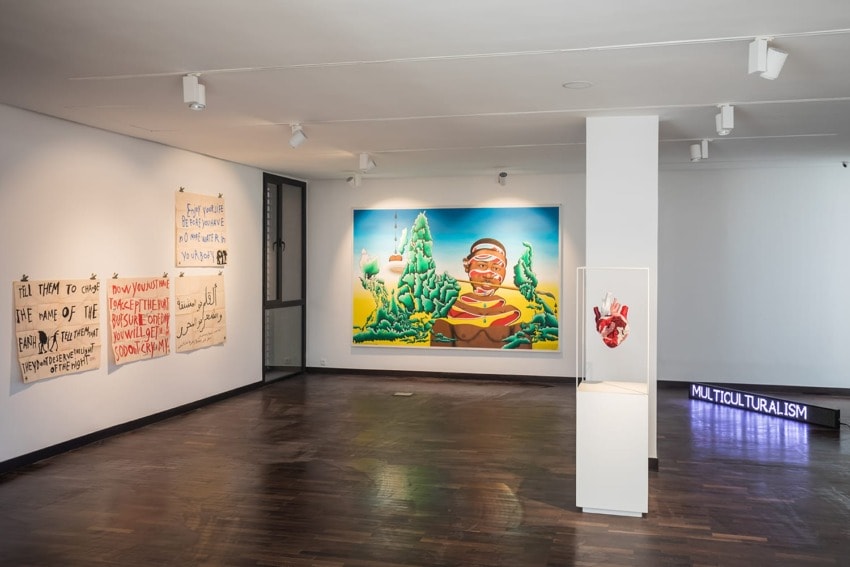 Art gallery interior with colorful paintings on walls, illuminated artwork titled "Multiculturalism," and a sculpture display.