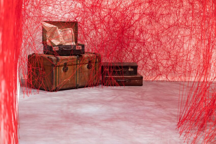Art installation with red threads surrounding vintage suitcases, creating an intricate web on a concrete floor.