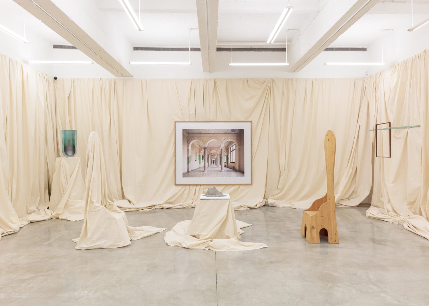 Art installation with draped beige fabric, wooden chair, covered objects, and a framed artwork on a concrete floor.