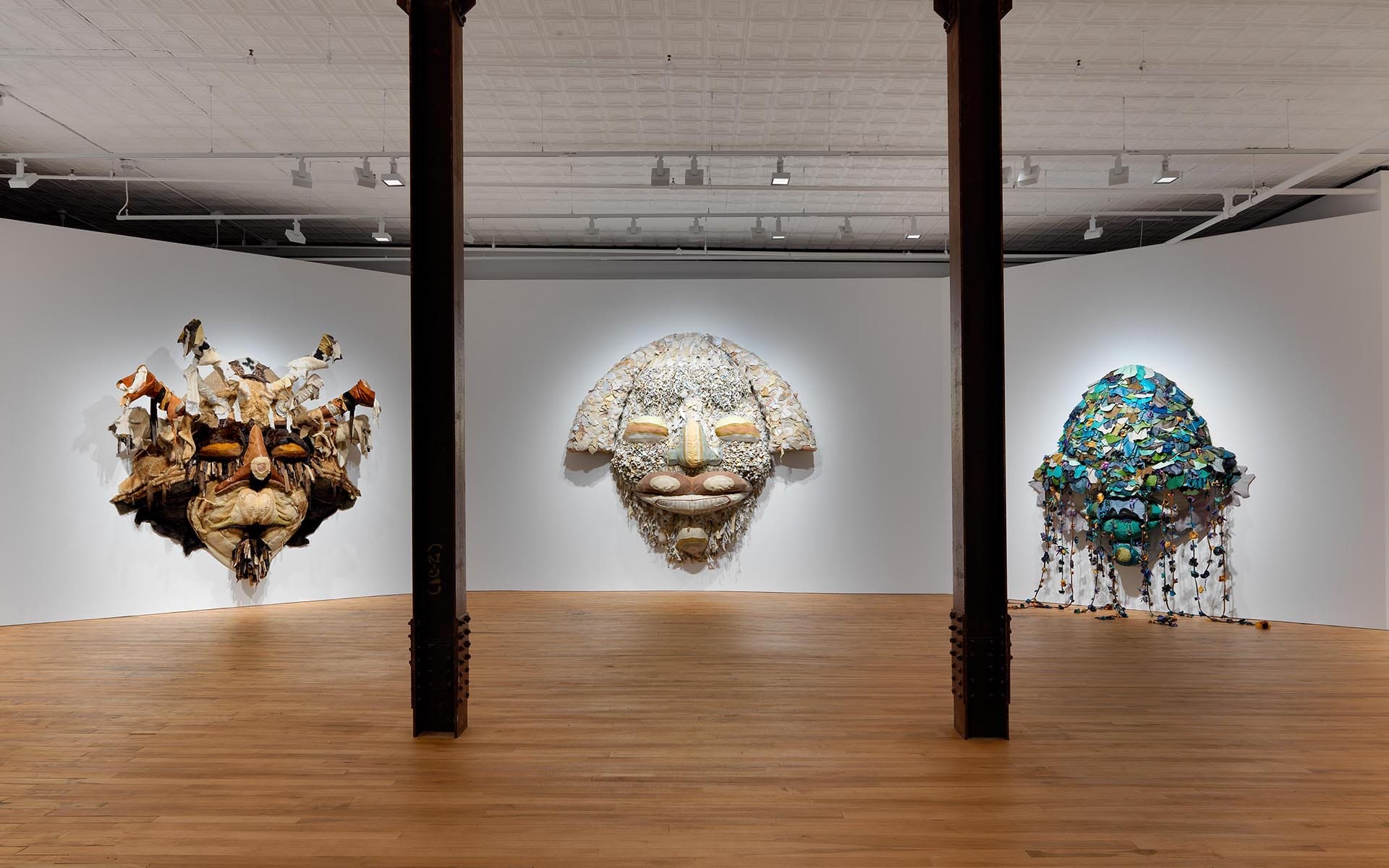 Art gallery featuring three large, colorful mask sculptures mounted on a white wall in a spacious room with wooden floors.