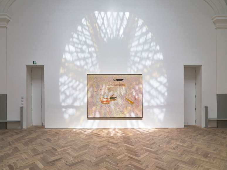 Large modern art piece displayed in a gallery with wooden floor and a patterned light reflection on the wall above the artwork.