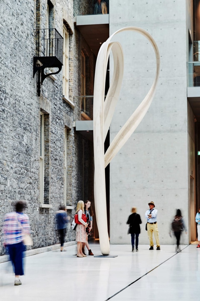 people walking past large modern wooden sculpture in art gallery with stone and concrete walls