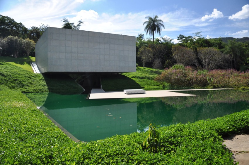 Modern concrete building over a reflective pond surrounded by lush greenery and a clear blue sky in the background.