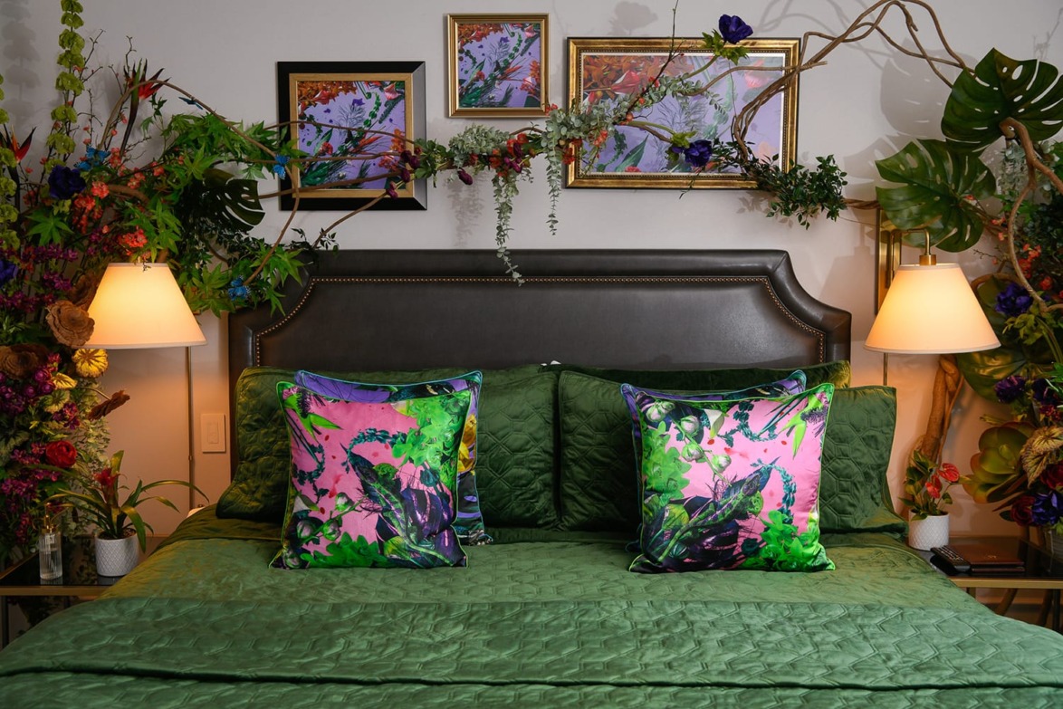 Luxurious bedroom with a green bedspread, vibrant floral pillows, decorative plants, and framed artwork above the headboard.