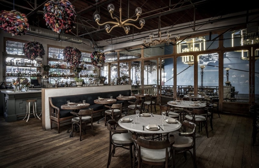 Vintage-style restaurant interior with wooden floors, round tables, elegant chandeliers, and vibrant hanging decorations.