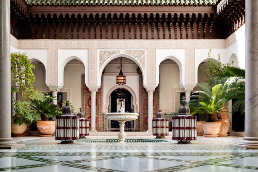 Elegant Moroccan courtyard with arched doorways, central fountain, lush greenery, and intricate architectural details.